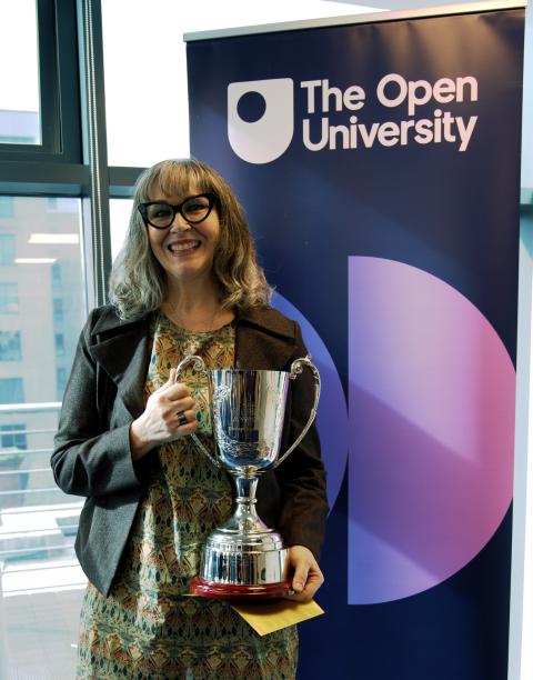  A woman with glasses and shoulder-length grey hair smiles while holding a large silver trophy. She is standing in front of a banner with "The Open University" logo. She wears a patterned dress and a brown jacket, appearing proud of her achievement. The background shows large windows with a cityscape visible outside.