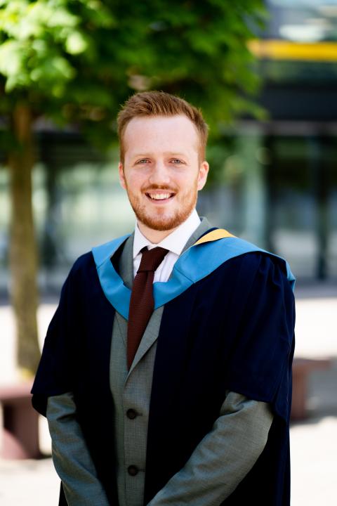 a man with red hair wears his graduation robes and smiles into the camera