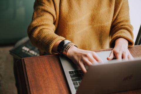 a woman sitting at a table at a laptop wearing a mustard jumper