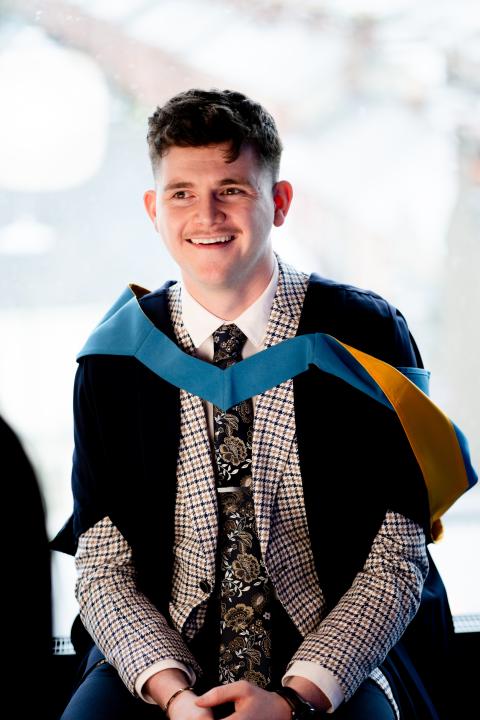 Odrhan sat down and dressed in OU graduation robes, smiling to someone just off centre left of the camera.
