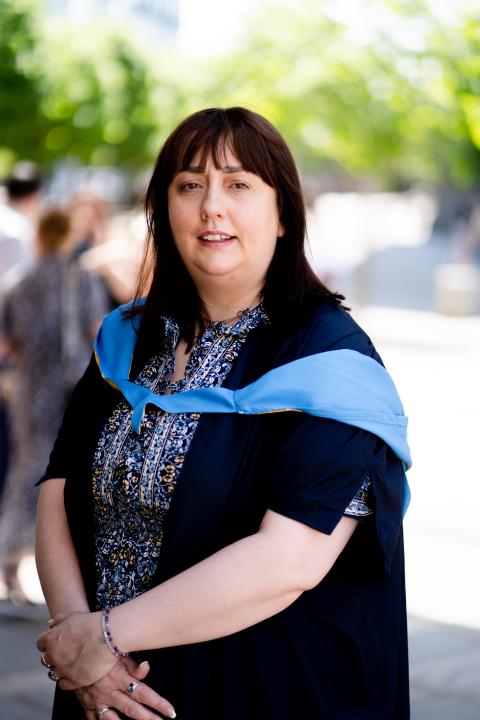 a woman with dark hair wears her graduation robes and smiles into the camera