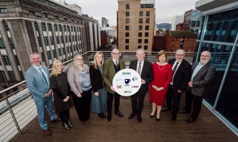  "Group of nine people standing outdoors on a rooftop terrace in an urban setting, holding a circular sign featuring various logos, including The Open University and other institutions. The individuals are dressed in business attire, smiling, with buildings and a cityscape visible in the background."