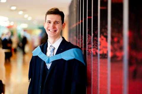 Rainers, in graduation robes, smiling to camera in a long hallway.