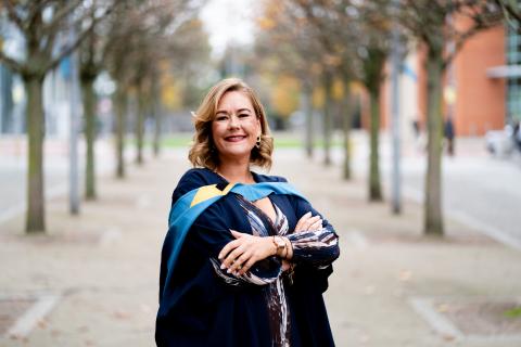 Sarahjane Dickson stands outdoors on a tree-lined path wearing an academic gown and hood, smiling with her arms folded as she celebrates graduating from The Open University.