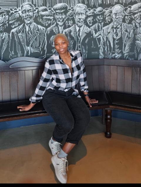 A person with short blonde hair sits casually on a wooden bench against a mural depicting a group of people in monochrome line art. They are wearing a black-and-white checkered shirt, black jeans, and white trainers, with one leg crossed over the other and a relaxed expression.