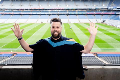 Sean standing with both hands in the air and a grin across his face. He's wearing OU degree robes and in the background you can see the pitch from Croke Park.