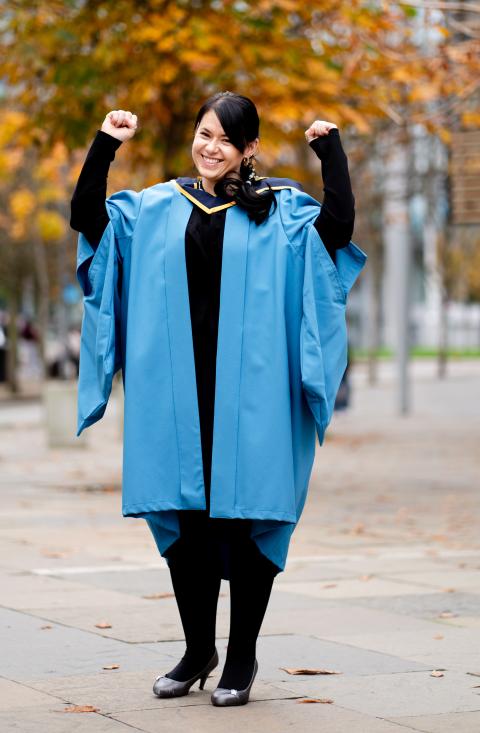 Supriya in OU graduation robes stood outside with her hands in the air celebrating.