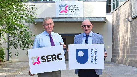 two men stand holding boards that have the SERC logo and The Open University on them