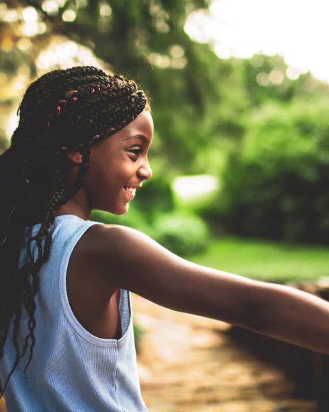 A smiling young girl with braided hair stands outdoors on a sunlit path, stretching her arms out as she walks beside a wooden railing, with trees and greenery softly blurred in the background.