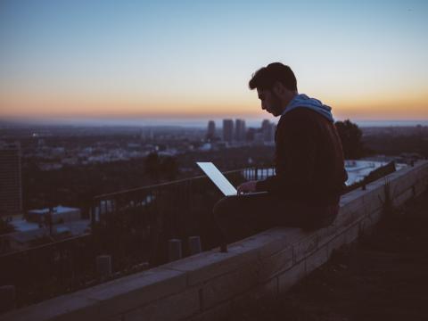 a young man sits on a wall at night using a laptop