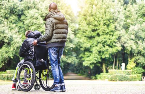 a man pushes another person in a wheelchair, they are wearing coats and are in a park