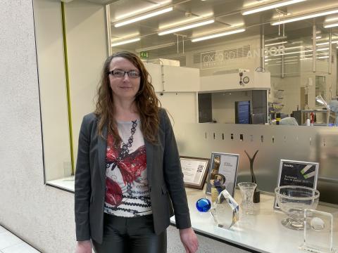 Tanya, a woman with long curly hair and glasses, stands indoors in front of a large window overlooking a lab with machines. Behind the window is also a low shelf displaying various awards.