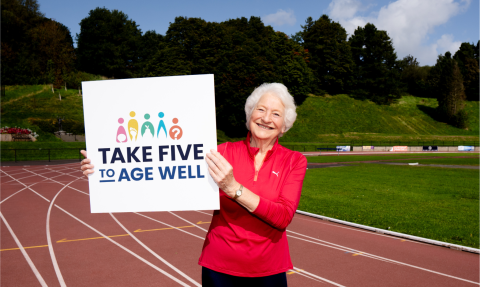 Mary Peters standing holding a take five to age well board whilst on a running track