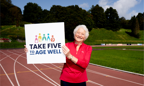 An elderly woman with white hair, wearing a red Puma sports top, stands smiling on a running track while holding a large sign that reads "Take Five to Age Well." The sign features colorful icons representing wellbeing. In the background, there is a green hillside, trees, and a bright blue sky with some clouds.
