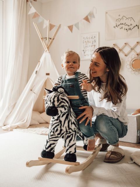 A mother crouches beside her toddler, who is seated on a zebra-patterned rocking horse. The child, wearing striped overalls and a shirt, looks at the camera, while the mother smiles warmly at them. The room is softly lit, decorated with a teepee, bunting, and wall art, creating a cosy, playful atmosphere.