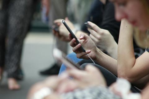 A close-up of multiple people sitting in a row, each engrossed in their smartphones. Their hands and devices are in focus, while their faces and backgrounds are blurred. The image captures a modern digital scene, likely in a public setting such as a waiting area or public transport.