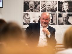 A man, dressed in a dark suit and white shirt, smiles while seated at an event. Behind him is a collage of black-and-white photographs of various individuals. The blurred heads of audience members are visible in the foreground, creating a sense of depth. The man appears engaged, possibly in a conversation or discussion. The setting suggests a formal or semi-formal event.
