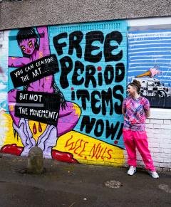 Person standing beside a colourful graffiti mural reading “Free Period Items Now” and “You can censor the art… but not the movement