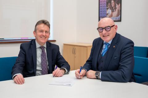“Two men in business attire sit at a table in an office, smiling as they sign a document with pens, suggesting a formal partnership or agreement.”