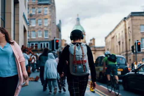 a young man walks along a street with his back to us carrying a back pack and with headphones on