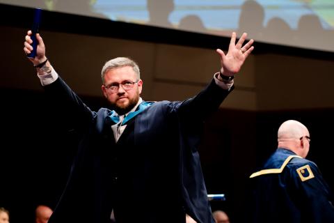 Peter is crossing the stage at a degree ceremony with his hands in the air in celebration.