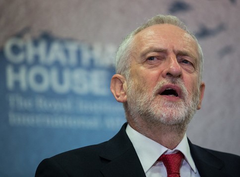 An image of Jeremy Corbyn, a man with grey-white hair and a white beard. He is wearing a black suit, white shirt and a red tie. He is speaking.