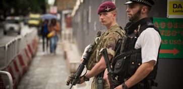 The image shows two men in the foreground. One is in camoflauge, with a red army hat and is holding a gun. The other is in a bulletproof vest, a police officer's hat, and is holding a gun.