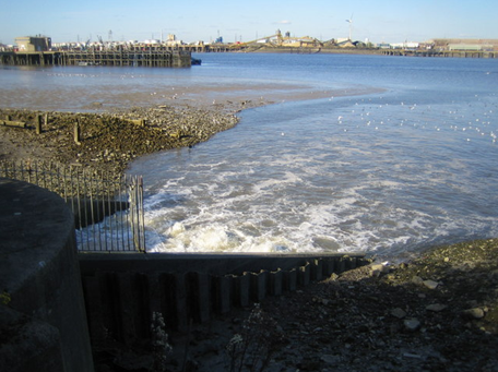The image shows water flowing into a large River. There are buildings in the background, and an opening in the foreground. Water is flowing from the opening.
