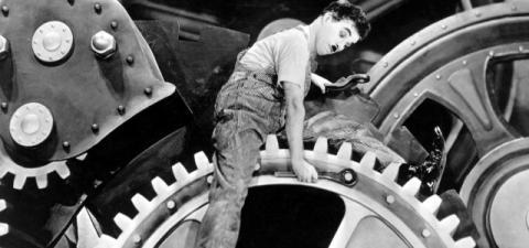 A man with a moustache (Charlie Chaplin) sitting on a gear holding a wrench.