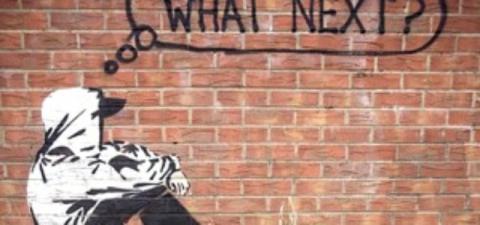 A brick wall with a picture of a person in a hoodie thinking 'What Next?'