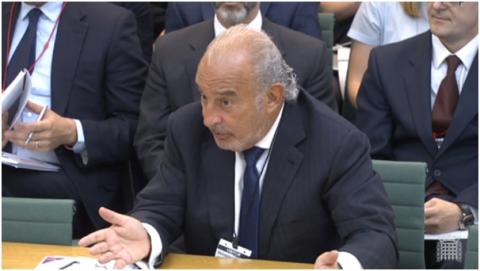 An older man in a suit with his arm outstretched in a questioning motion.