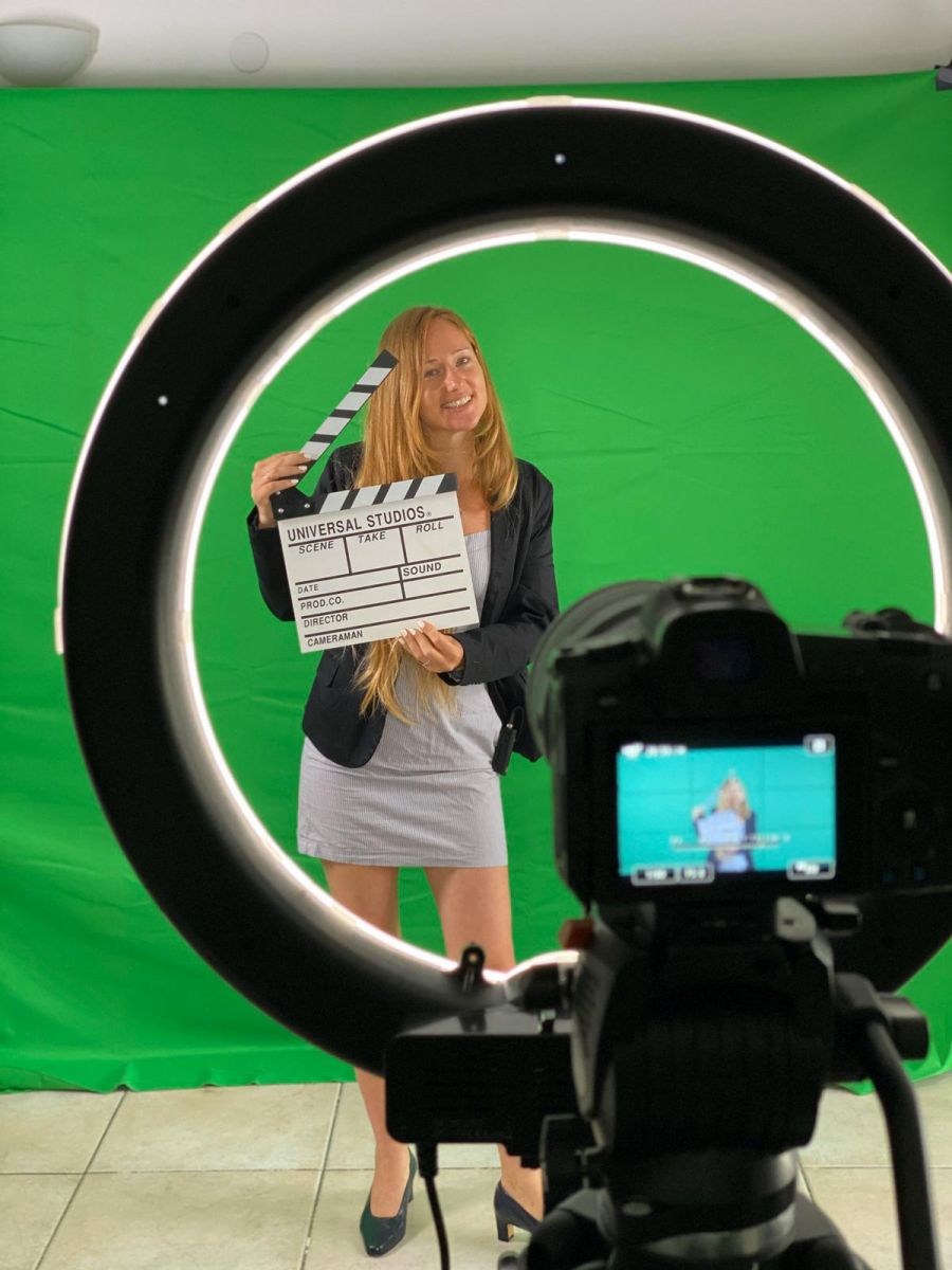 Bea Baharier in front of a green screen, holding a clapperboard, seen from behind a camera.