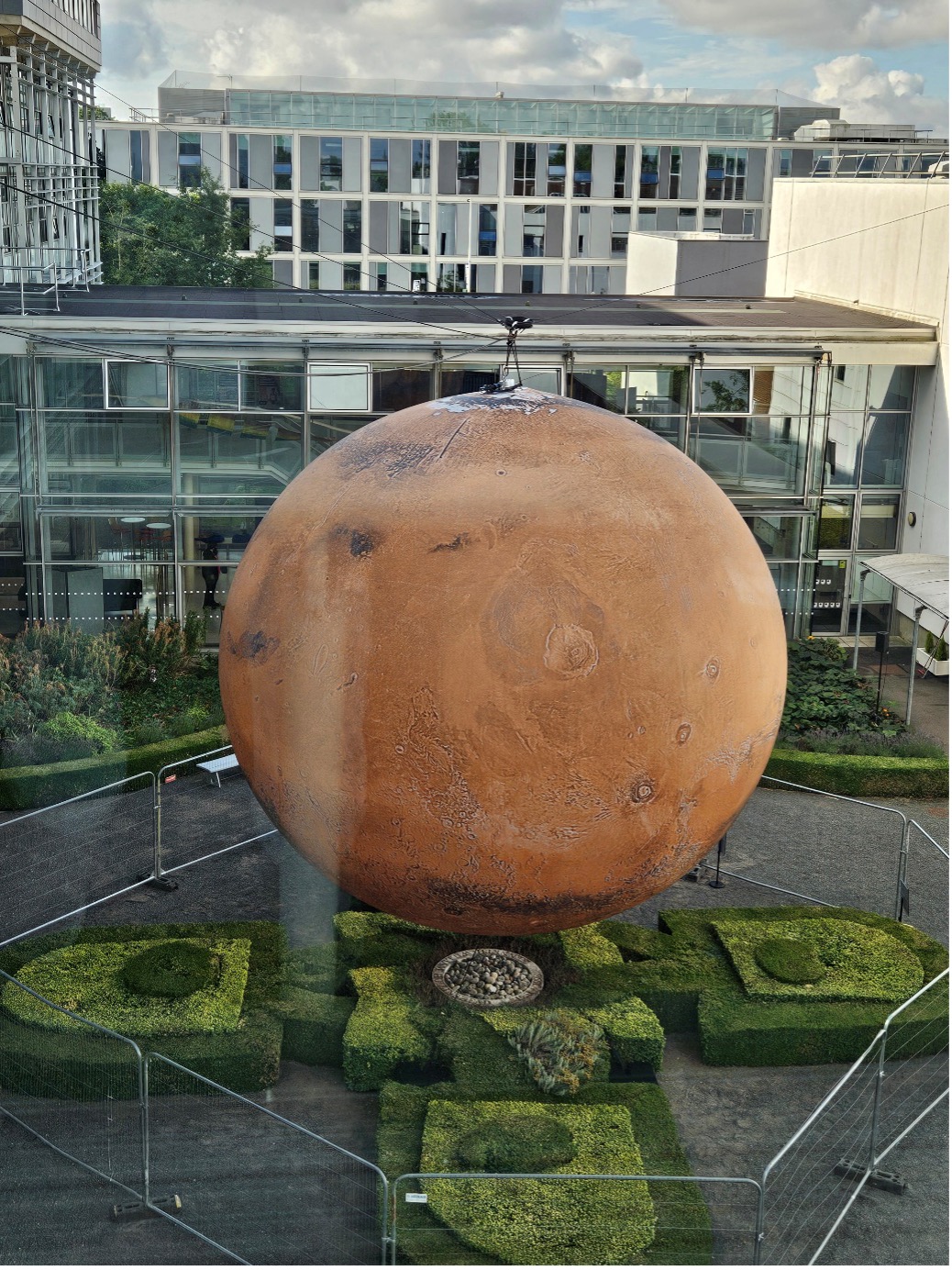 a large glove of Mars, suspended above a courtyard with manicured hedges, in front of a glass building