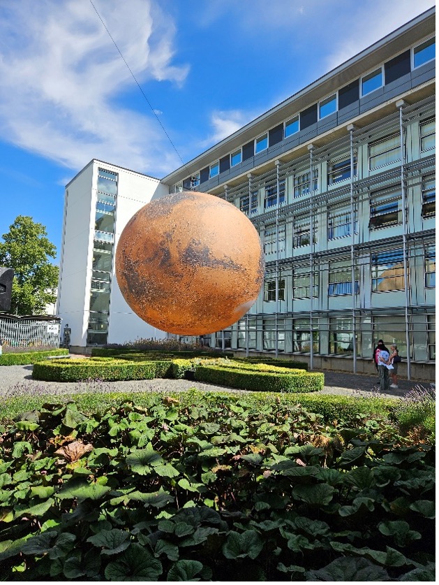 A large globe of Mars in front of a building with many glass windows with the focus on lavender plants in the foreground
