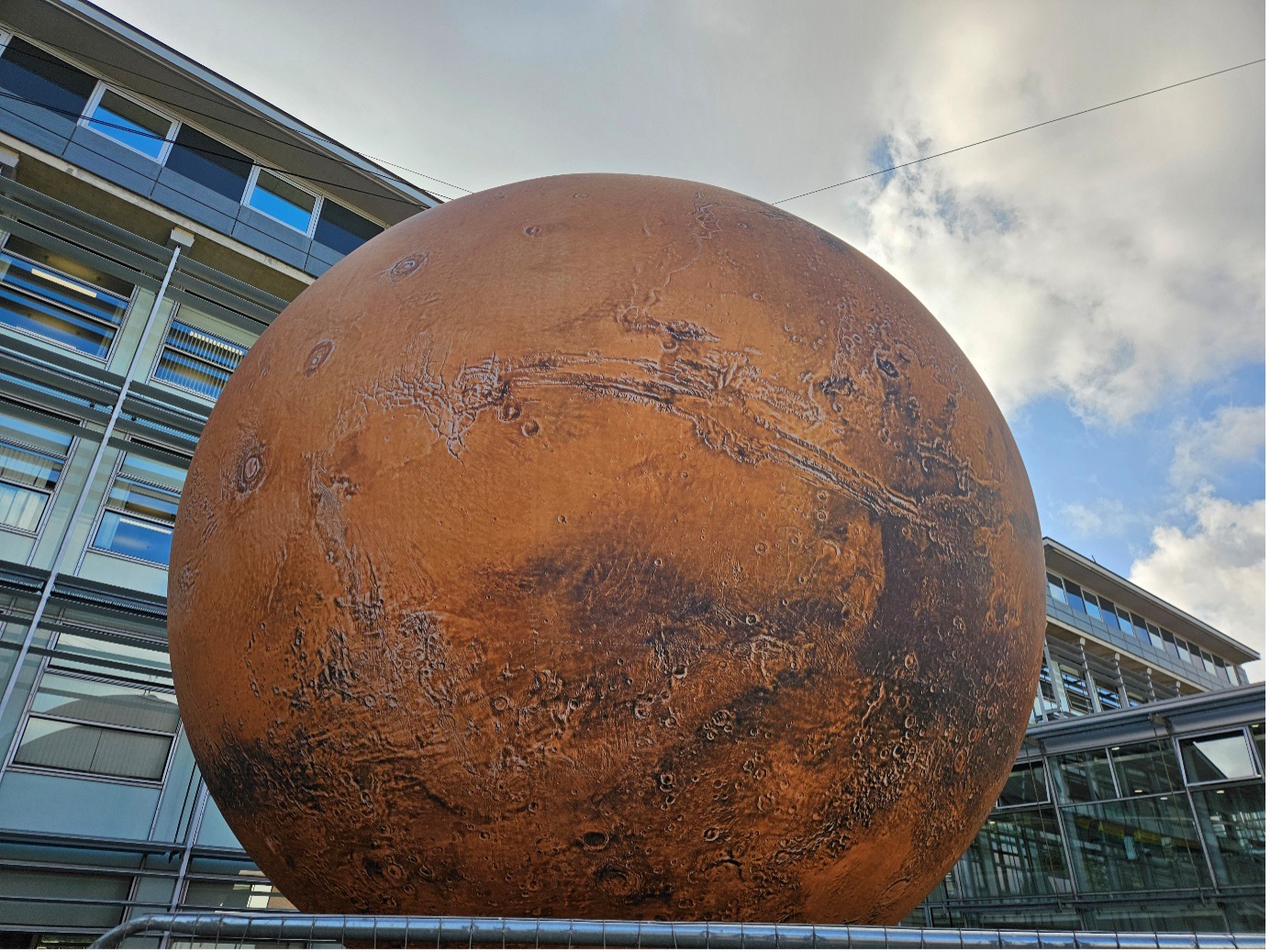 A large globe of Mars in front of a building with many glass windows with a cloudy sky behind
