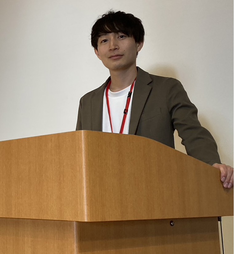 Shun Standing at a podium in a white t-shirt, brown blazer witha red lanyard. 
