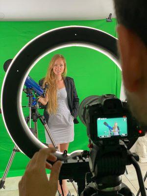 Bea Baharier standing in front of a green screen, next to a telescope. photographed through a light ring, behind which you can see the camera.