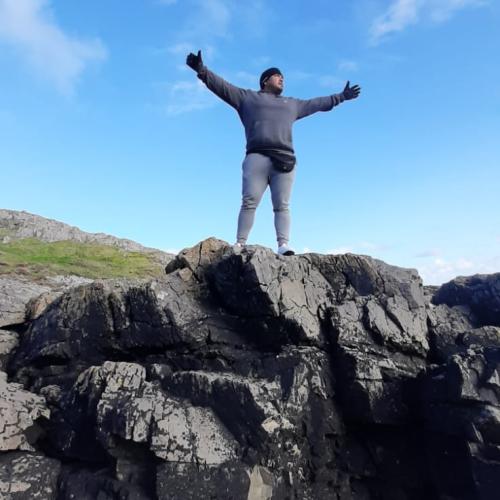 Armando stands on a high rock with his arms stretched wide with the blue expanse of the sky behind him
