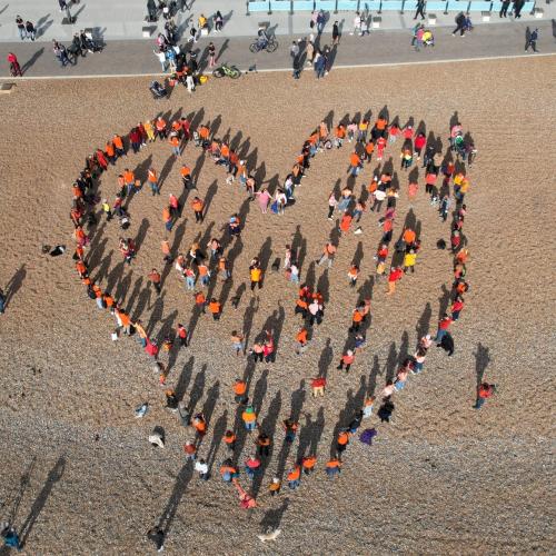 Community in Brighton stood on the beach in the shape of a heart, all wearing orange