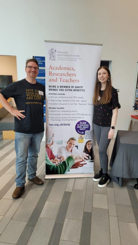 Two people standing and smiling. Thy are standing either side of a DARTP conference poster