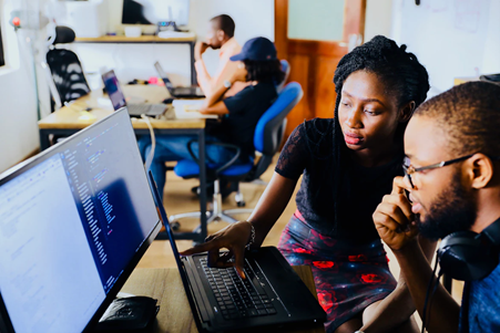 Two people sitting at a desk together, studying information on a computer screen. Two further people out of focus in the background