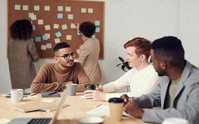 A small team of people talking at a table. In the background two other people are looking at sticky notes on a corkboard