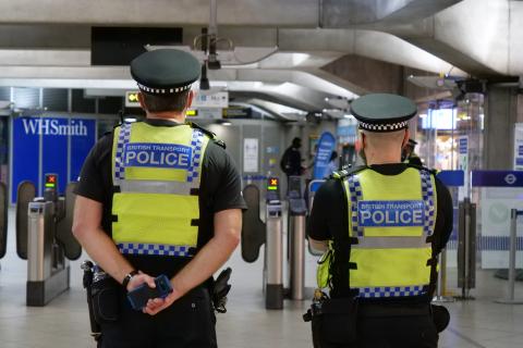 Two policeman in uniform standing in a train station. Their high visibility jackets have the word "Police" written on the back.