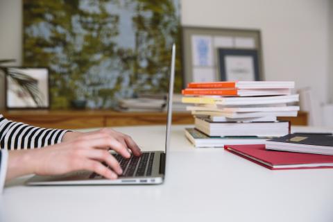 A person sitting at a desk with a pile of books, typing on a laptop