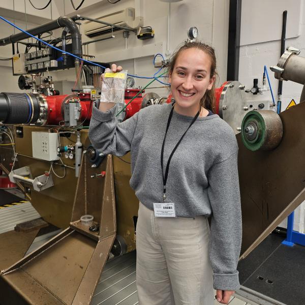 Student Kayleigh Moore standing in front of the All-Axis Light Gas Gun holding up one of her composite shield targets following a hypervelocity impact test.