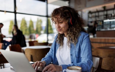 Person wearing headphones and facing an open laptop screen whilst sitting by a table in a cafe setting
