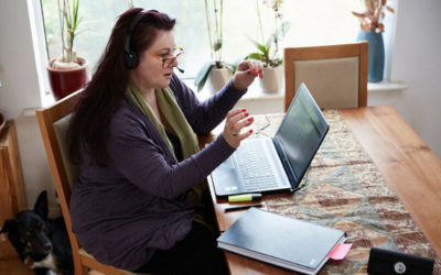 Person sitting at a kitchen table, wearing headphones and facing an open laptop screen