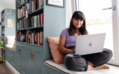 A woman working on her laptop whilst sitting at a window seat in her home