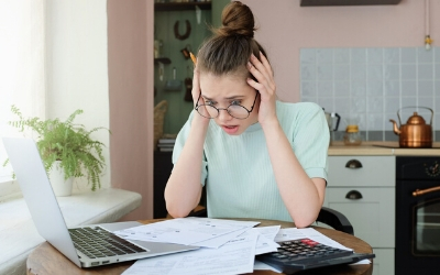 Woman with worried expression, sitting at kitchen table with papers, calculator and an open laptop