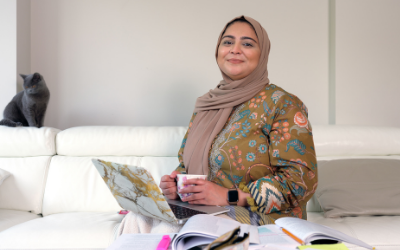 Student Halima, seated on a sofa with a laptop and holding a mug. A cat is nearby.